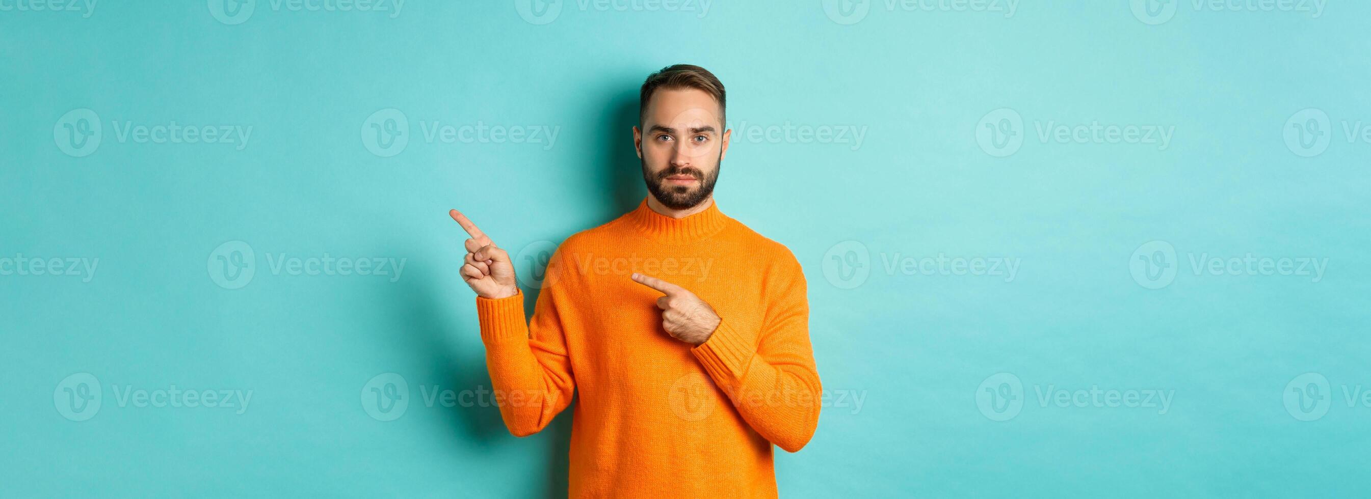 Handsome and serious bearded man in orange sweater pointing right, showing advertisement or logo, standing over turquoise background photo
