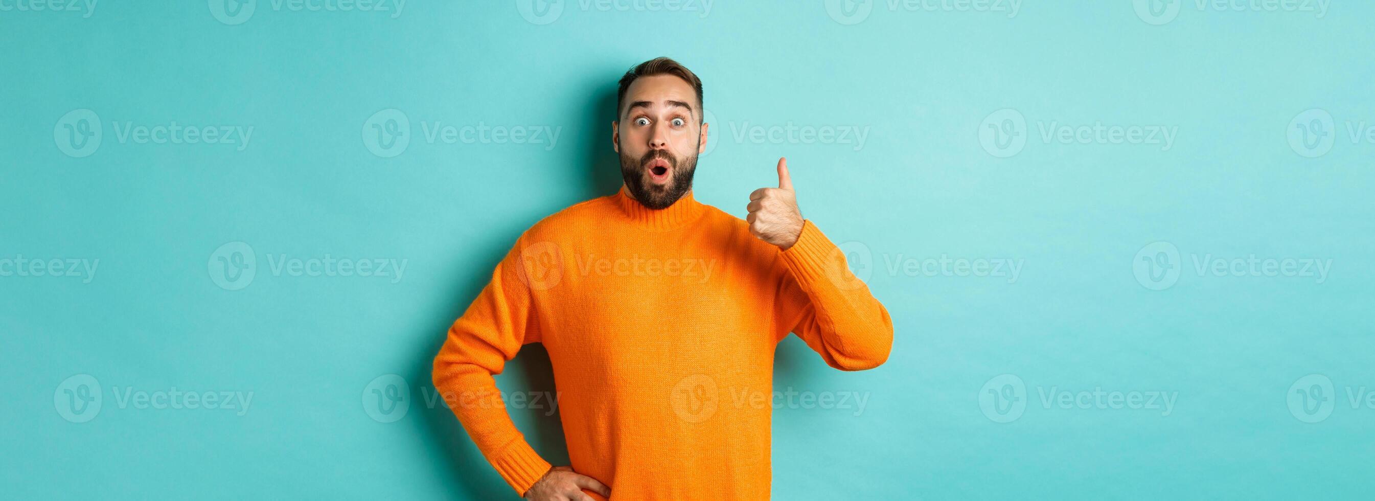 Well done. Handsome bearded man showing thumb up, praising good work, recommend excellent product, like and approve, smiling satisfied, standing over light blue background photo