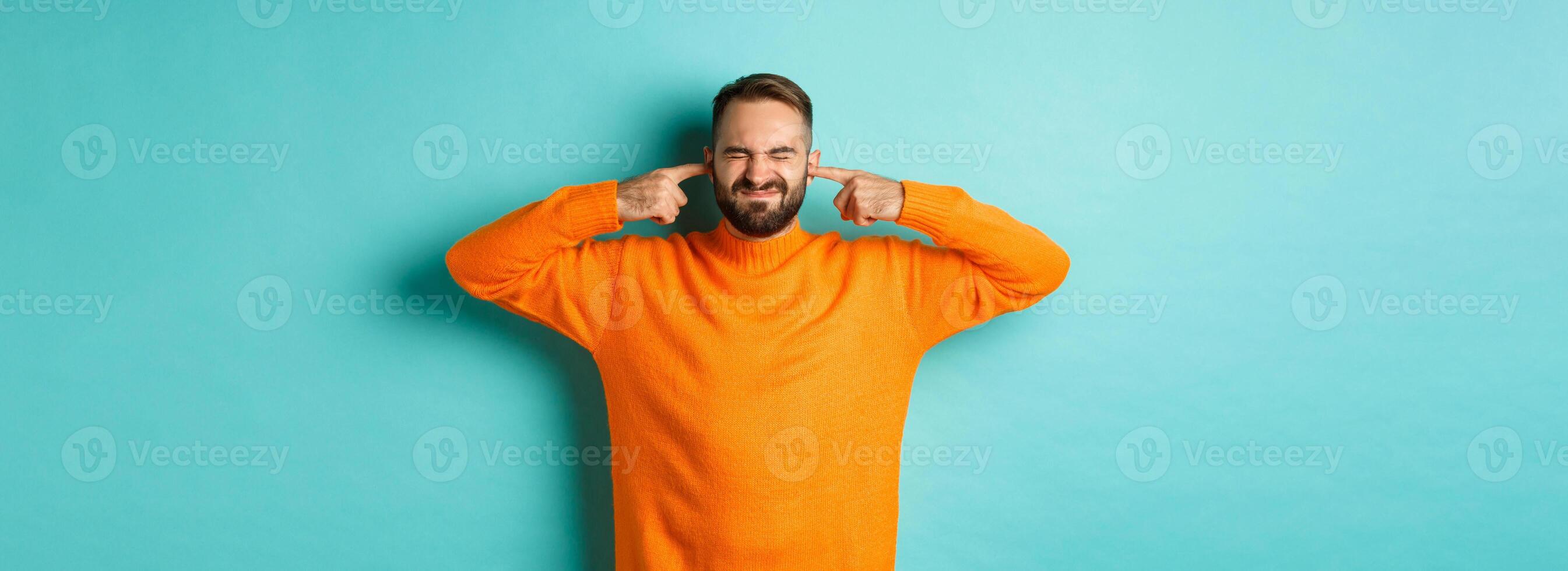 Displeased man shut ears and grimacing, loud neighbours, standing annoyed with noise, light blue background photo