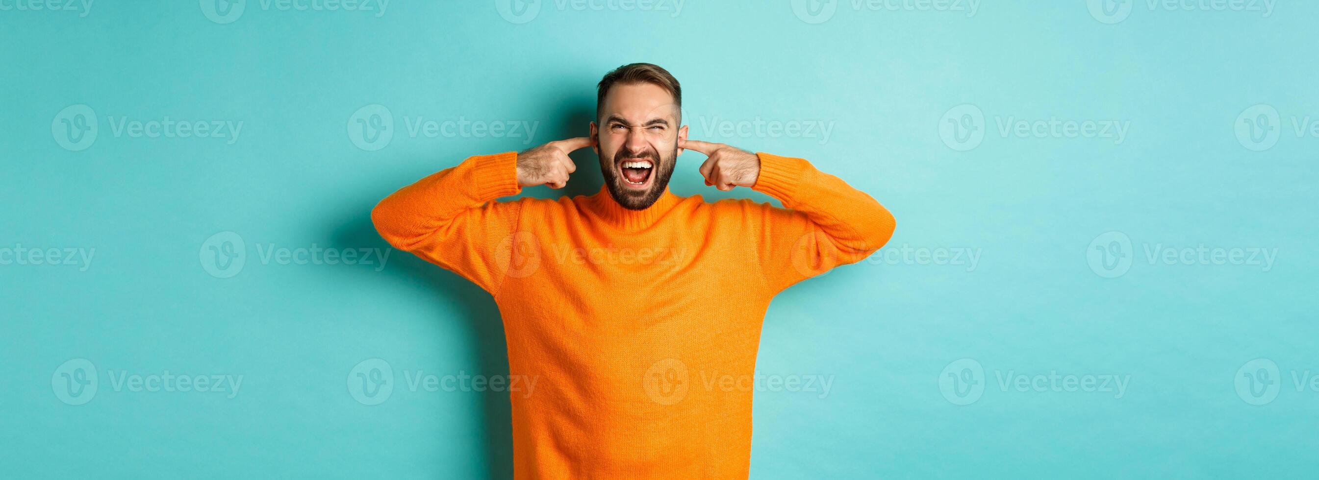 Displeased man shut ears and grimacing, loud neighbours, standing annoyed with noise, shouting angry and distressed, light blue background photo