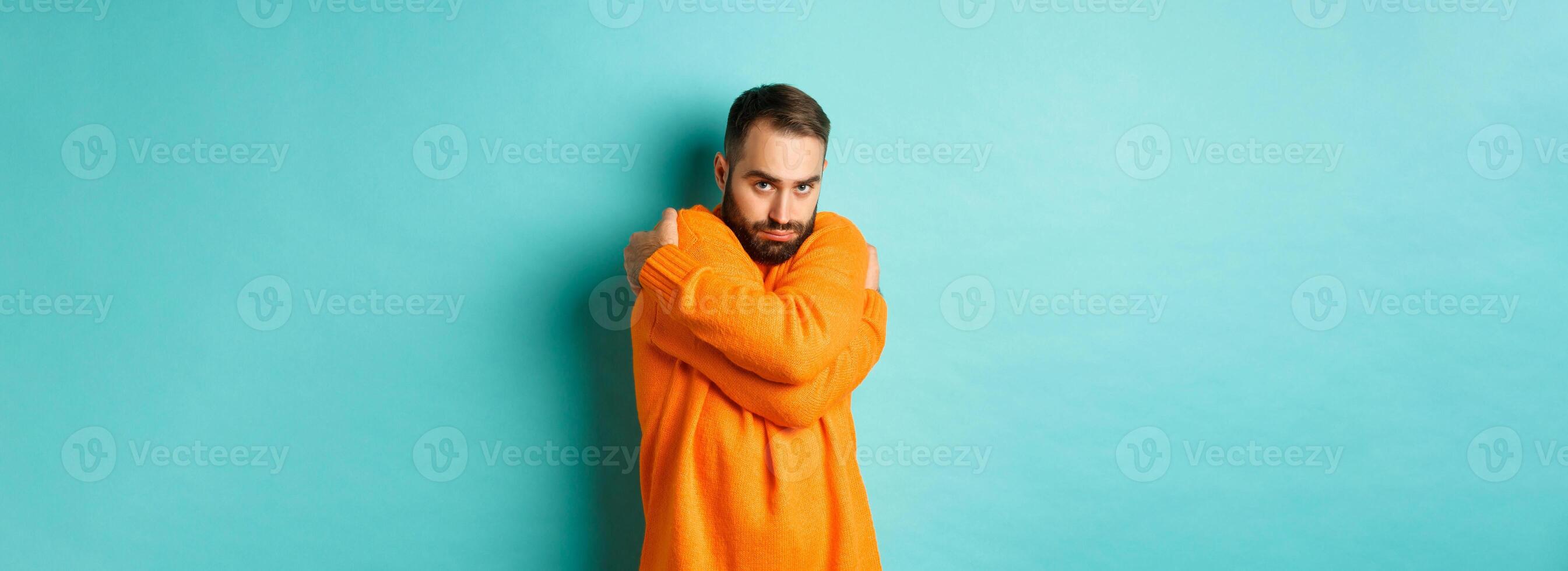 Timid man feeling offended and defensive, hugging himself and looking suspicious at camera, standing over light blue background photo