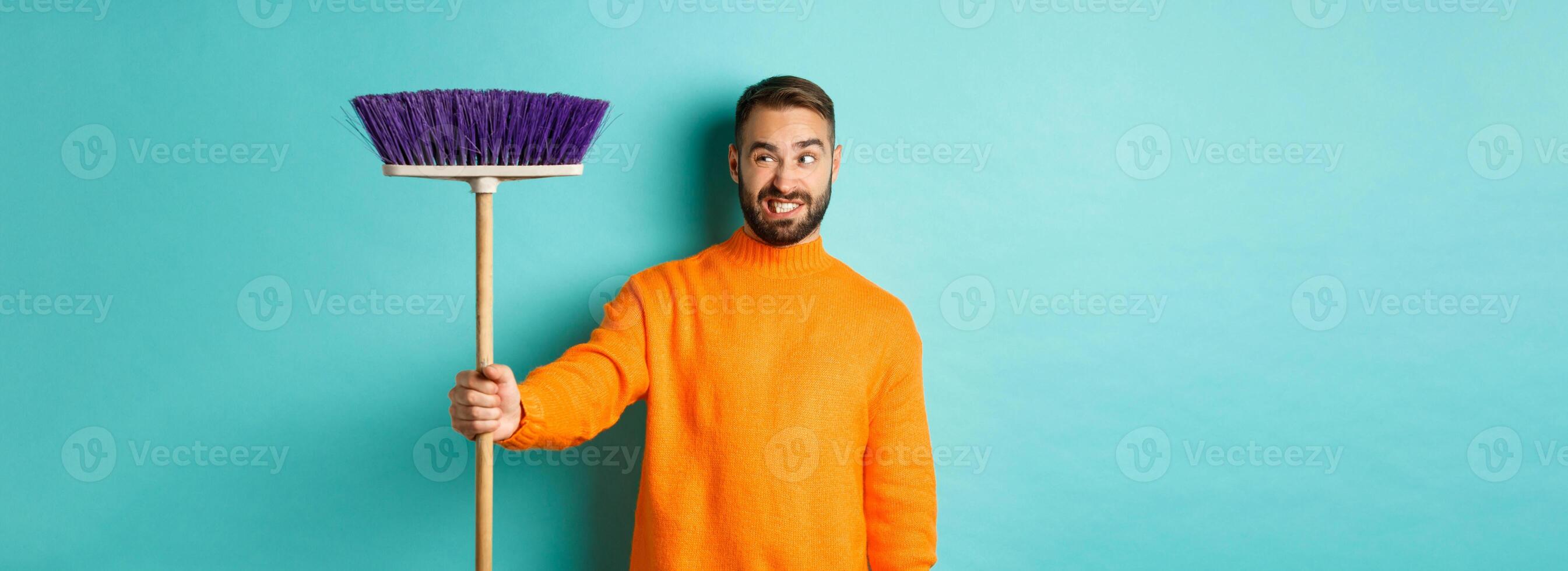 Boyfriend looking confused at broom, forced clean house, doing chores, standing displeased against light blue background photo