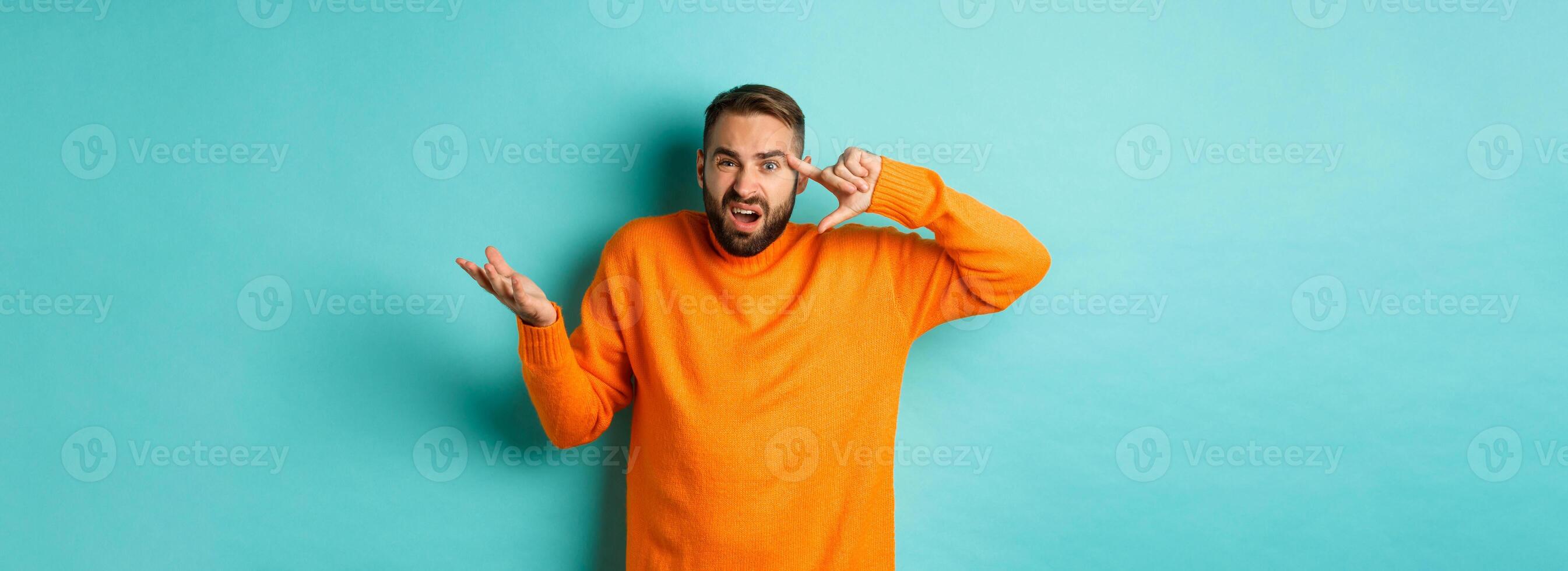 Angry man pointing at head, are you crazy gesture, scolding someone, standing over light blue background photo