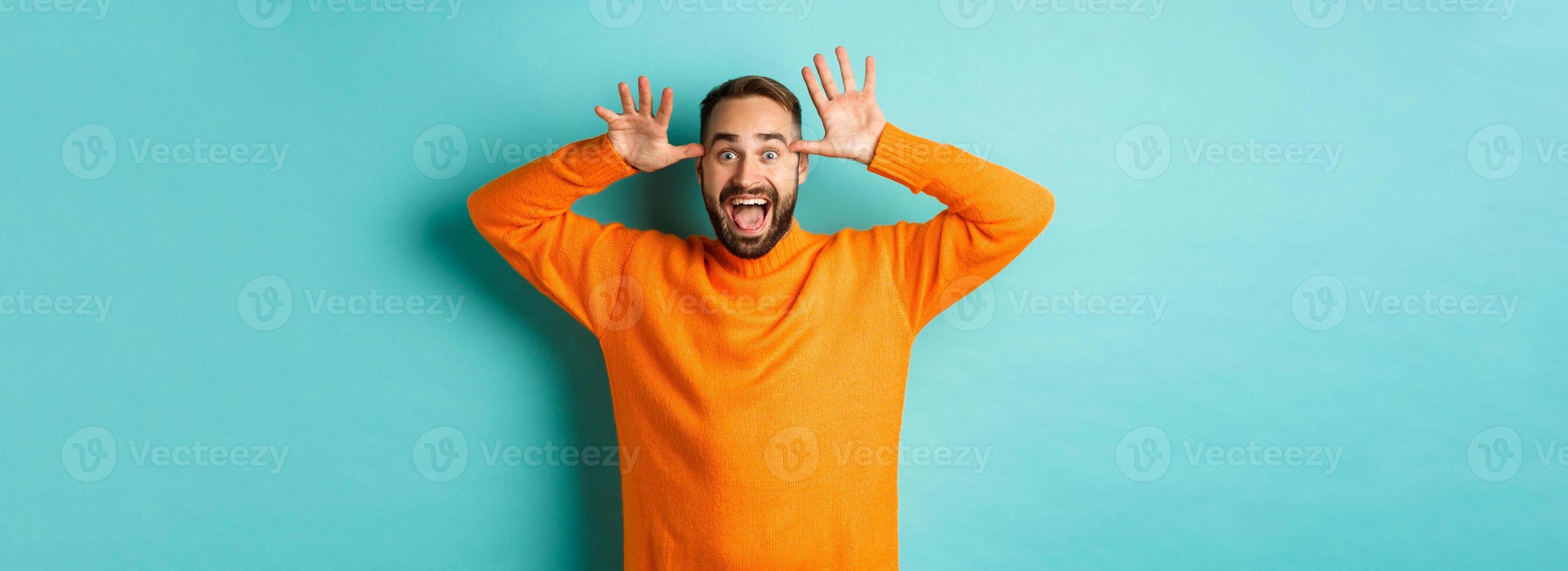 Image of handsome caucasian man making funny faces, mocking someone and smiling, standing against light blue background photo