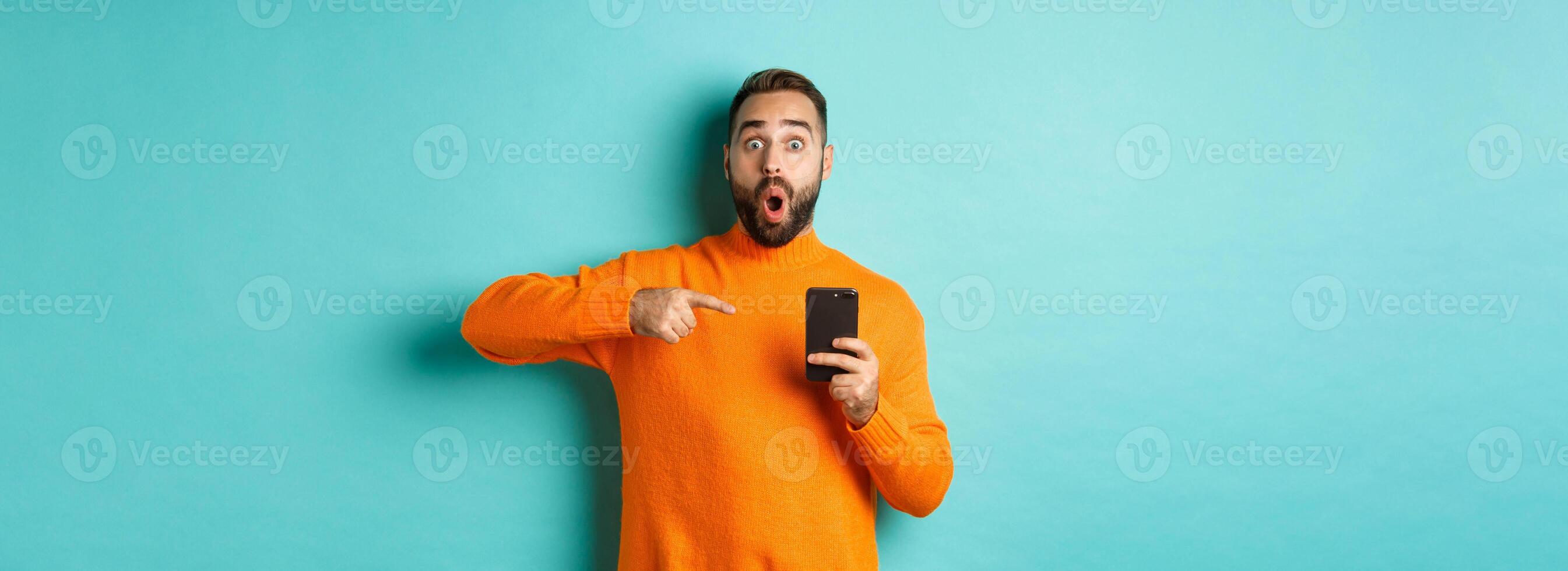 Image of impressed man showing something on mobile phone, pointing at smartphone and gasping amazed, standing in orange sweater over light blue background photo