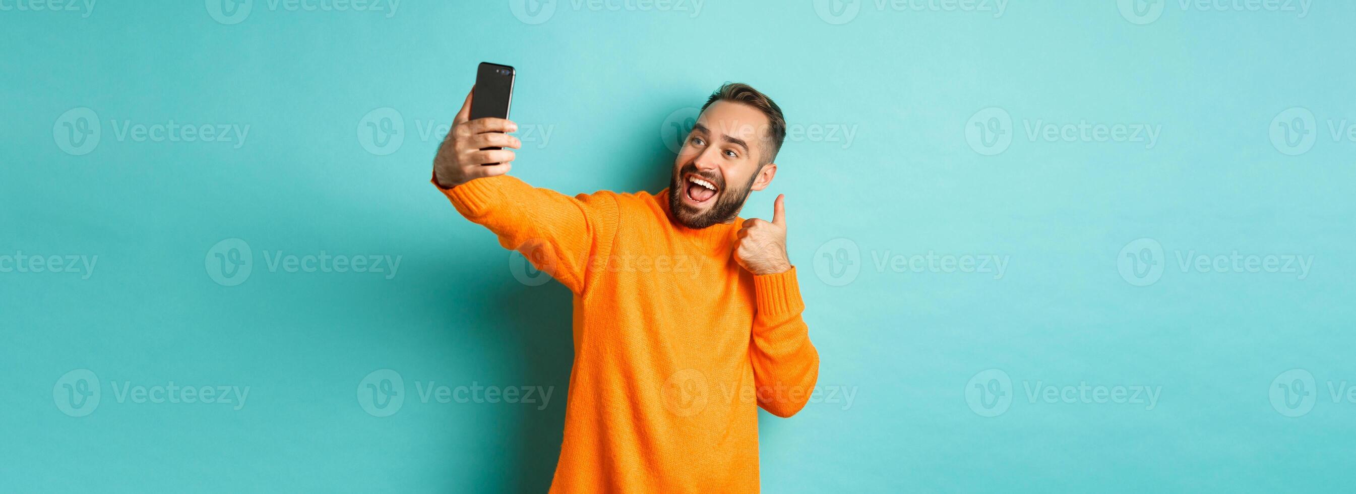 Handsome young man taking selfie on mobile phone, showing thumbs up at smartphone camera, recording vlogg, standing over light blue background photo