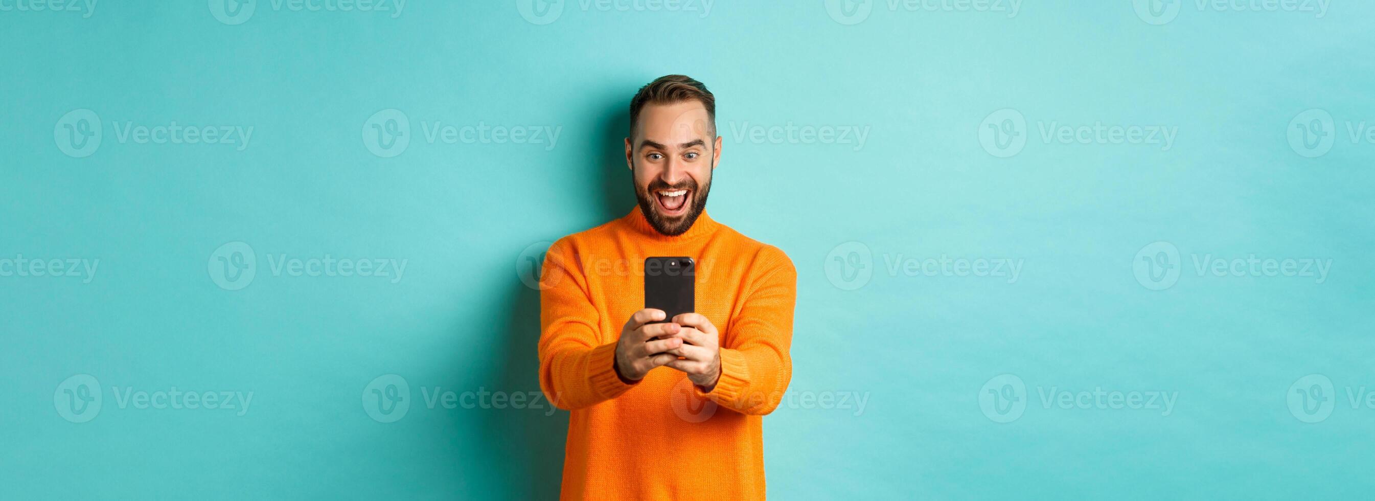 Image of man reading awesome message on mobile phone, looking at smartphone screen with amazement, standing over light blue background photo