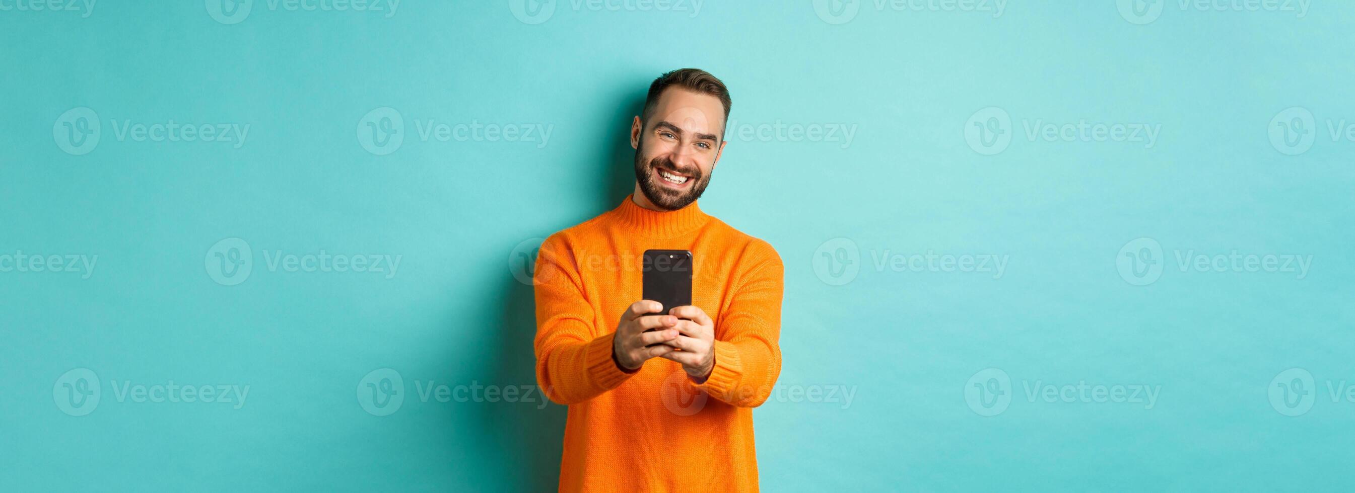 Happy handsome man taking photo on mobile phone, take pictures with smartphone camera, standing over light blue background