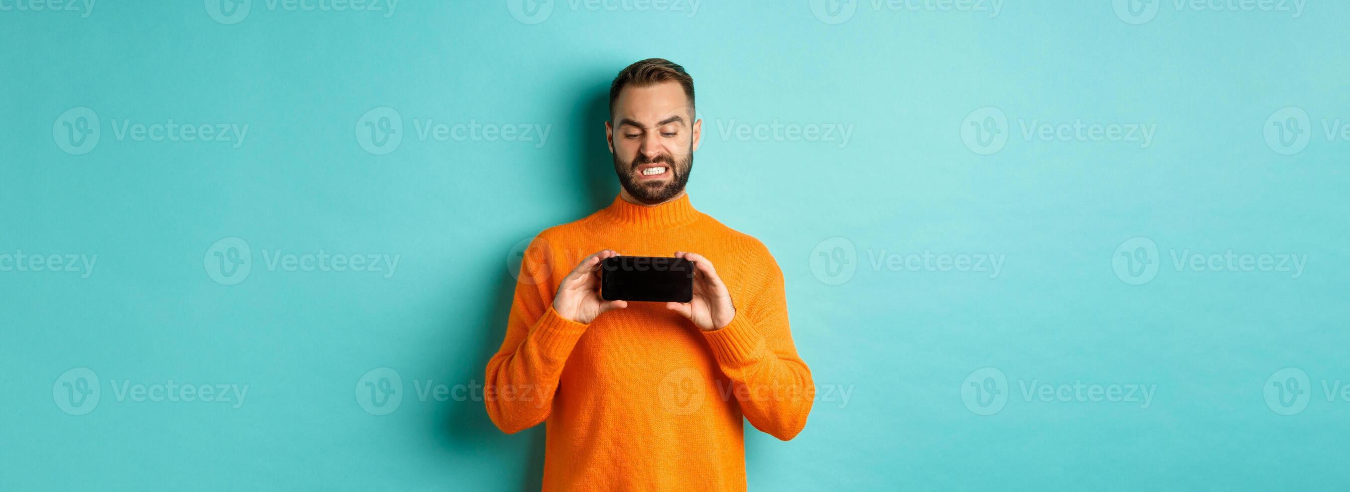 Image of man cringe at something on mobile screen, staring disgusted at display, showing smartphone, standing over light blue background photo