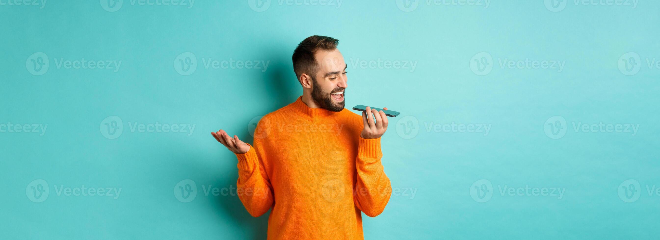 Happy man talking on speakerphone, gesturing and recording voice message on mobile phone, standing in orange sweater over light blue background photo