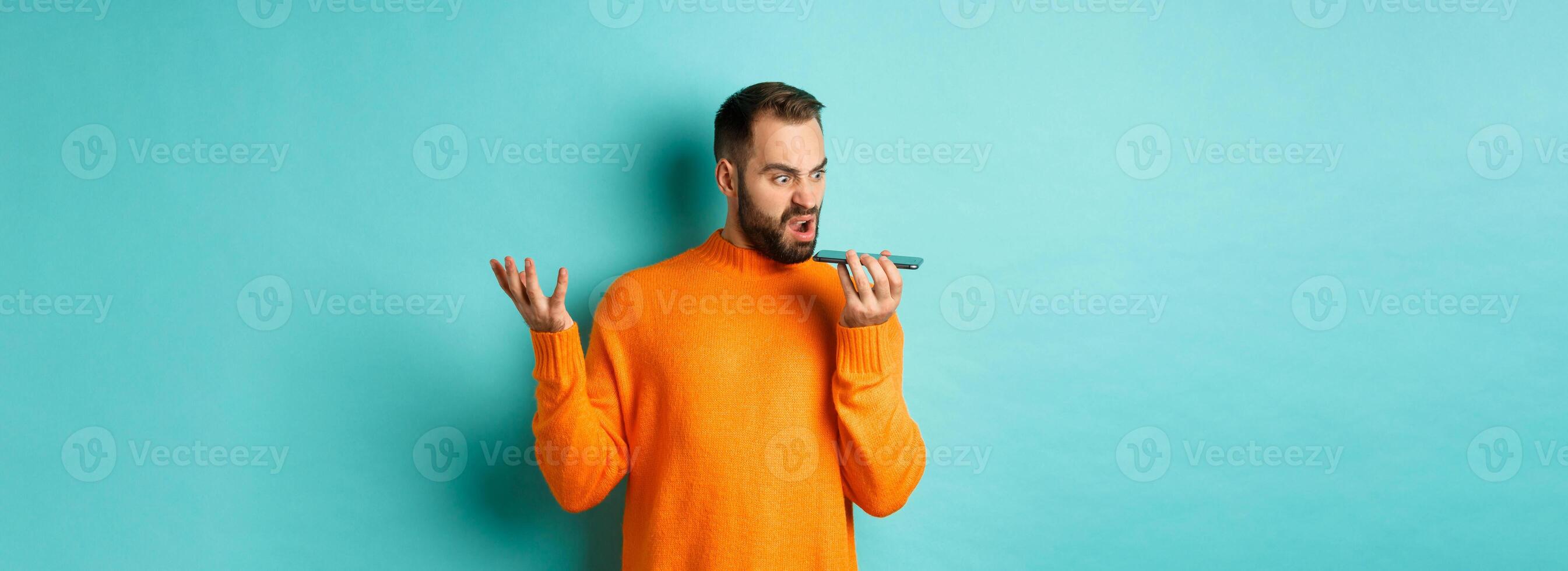 Angry man arguing on speakerphone, record voice message with mad face, standing over light blue background in orange sweater photo