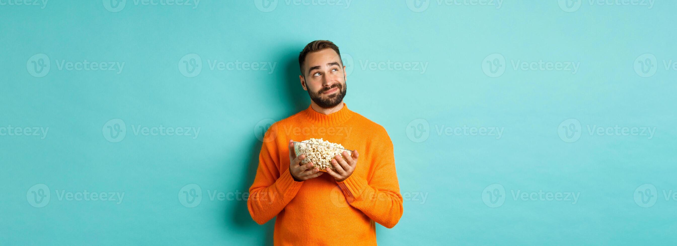Handsome young man in orange sweater, looking thoughtful at upper left corner, holding popcorn, picking movie, blue background photo