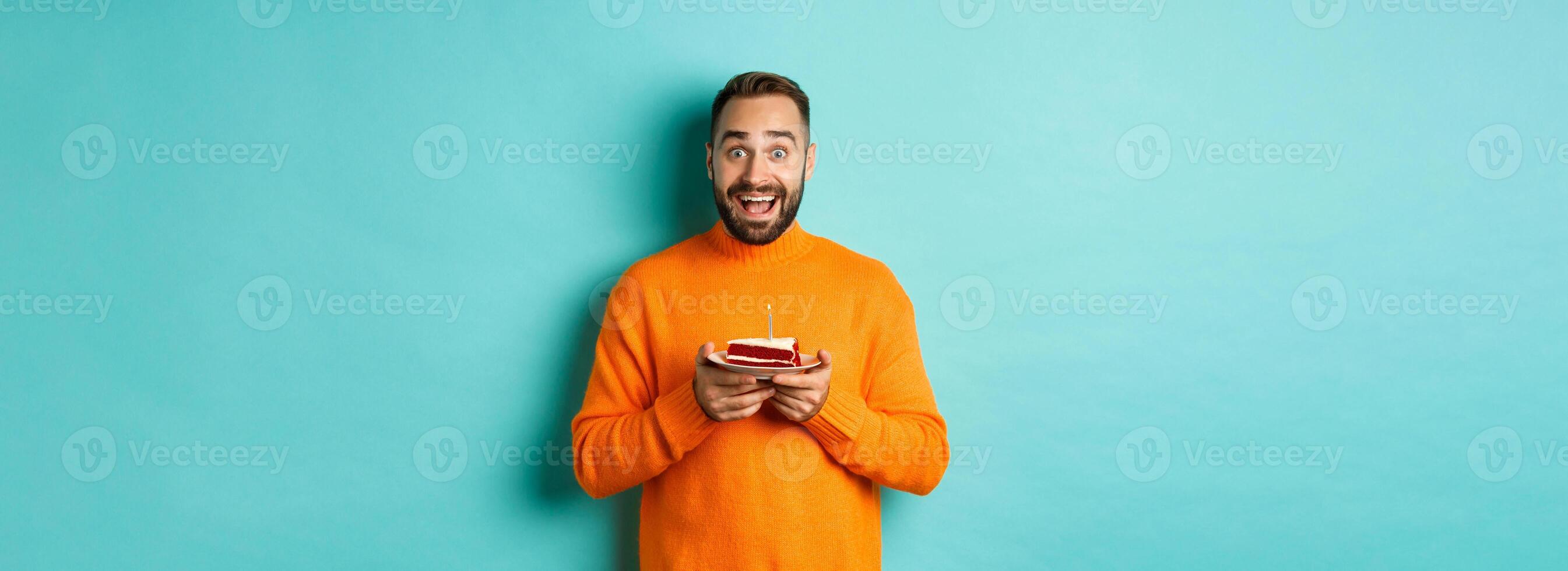 Happy adult man celebrating birthday, holding bday cake with candle and smiling, standing against turquoise background photo