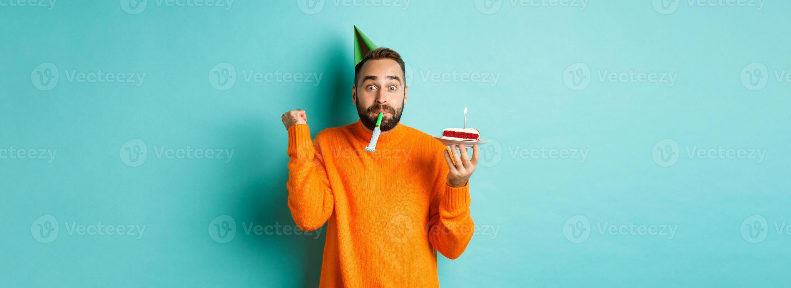 Happy birthday guy celebrating, wearing party hat, blowing wistle and holding bday cake and doing fist pump excited, standing over turquoise background photo