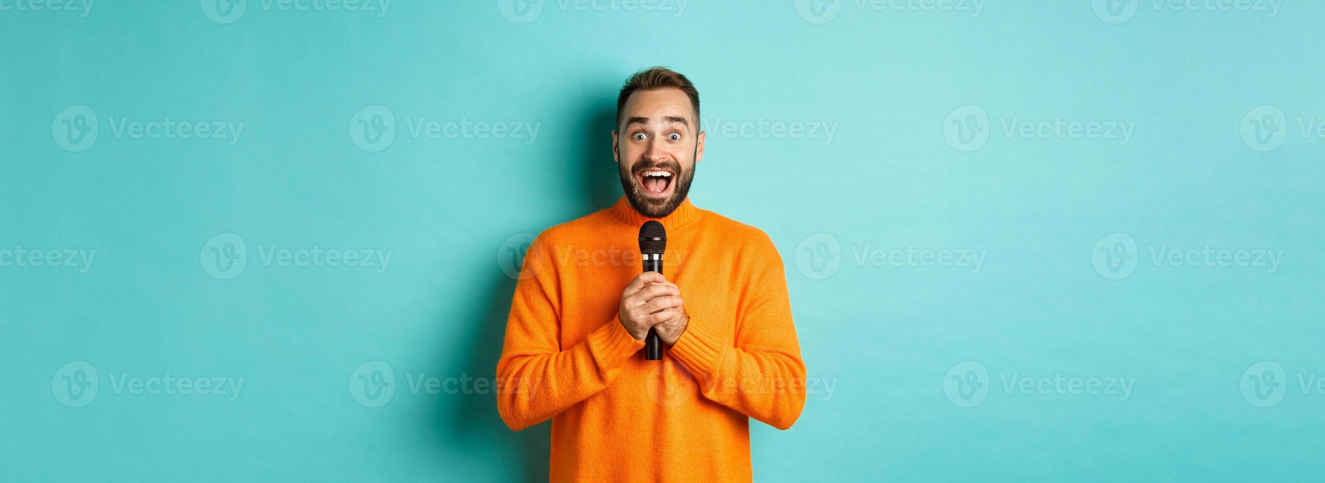 Happy adult man singing karaoke, holding microphone and looking at camera, standing in orange sweater against turquoise background photo