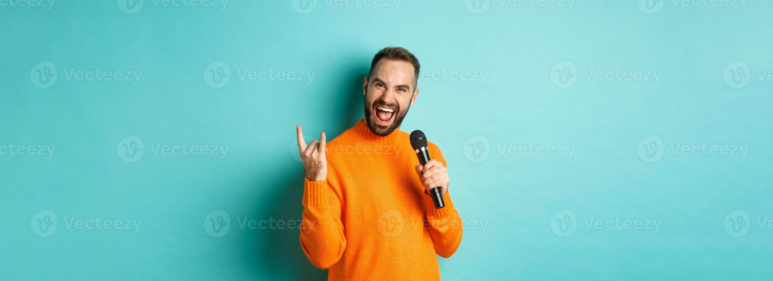 Joyful handsome man singing song in microphone, showing rock-n-roll gesture excited, standing over turquoise background photo