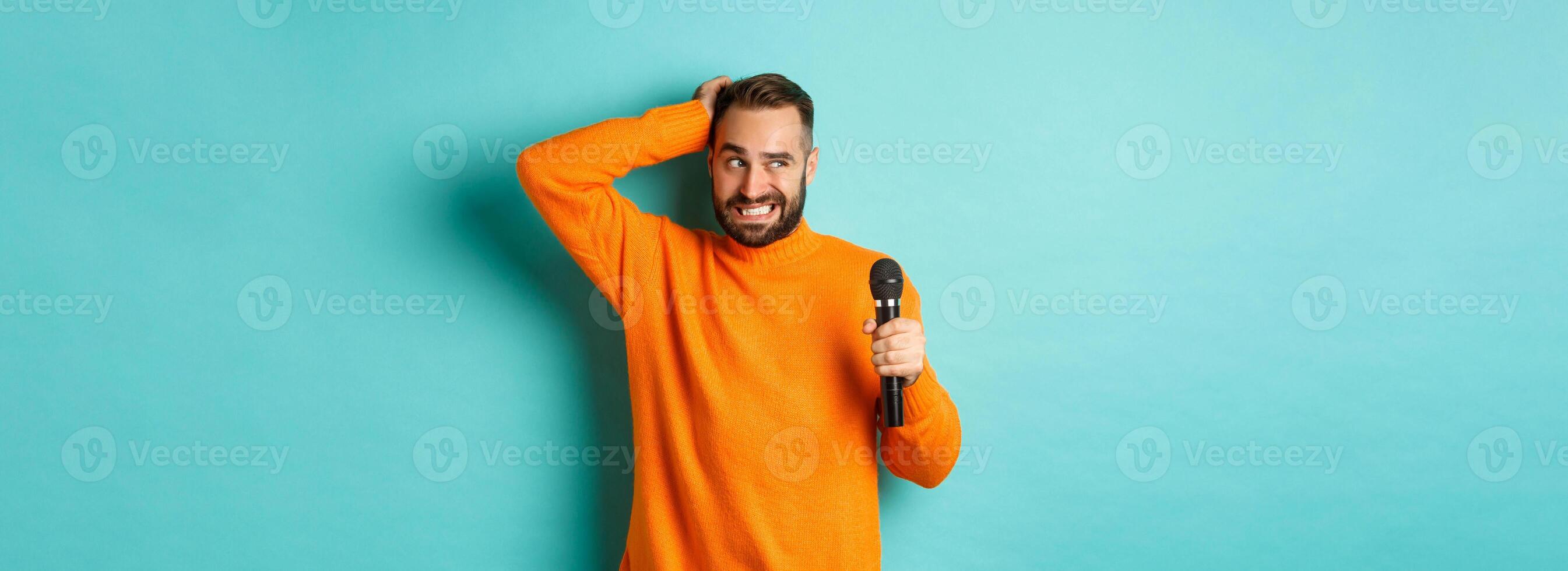 Shy and awkward adult man scratching head, holding microphone before singing, standing over blue background photo