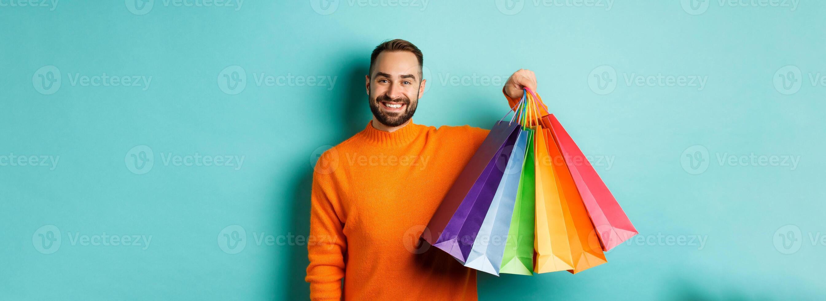 Happy handsome man holding shopping bags and smiling, buying presents, standing over turquoise background photo