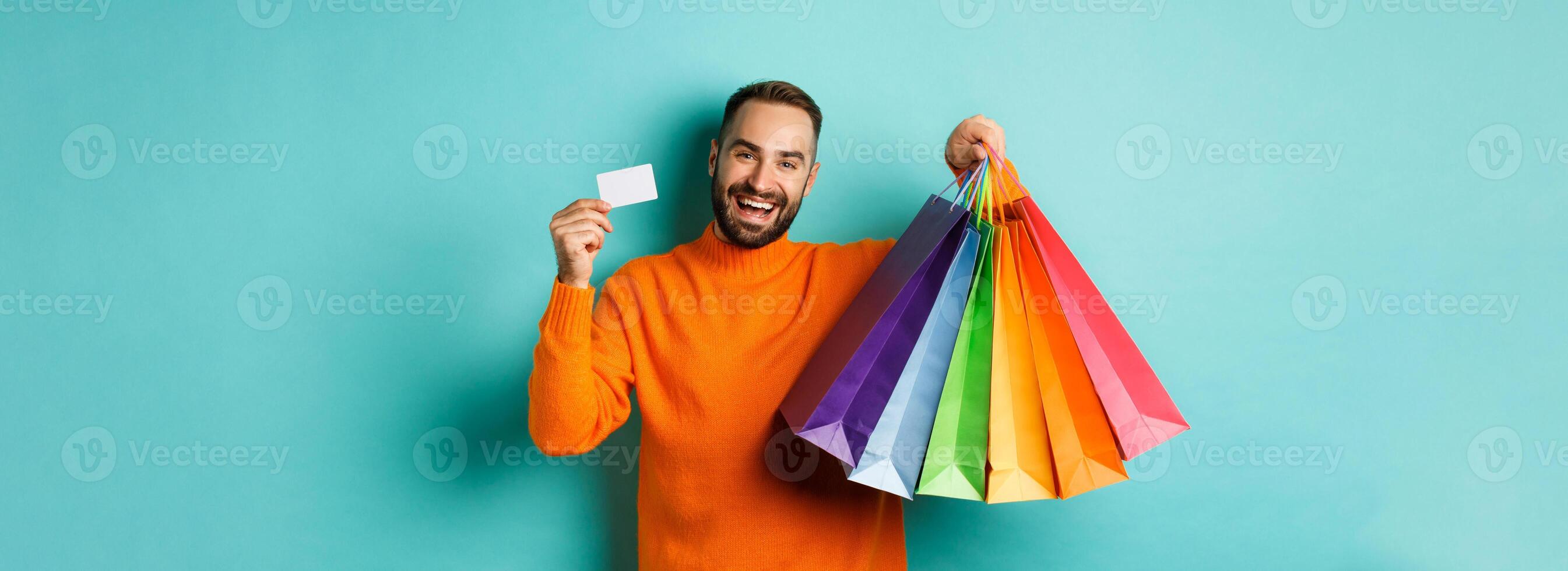 Happy aduly man showing credit card and shopping bags, standing against turquoise background photo