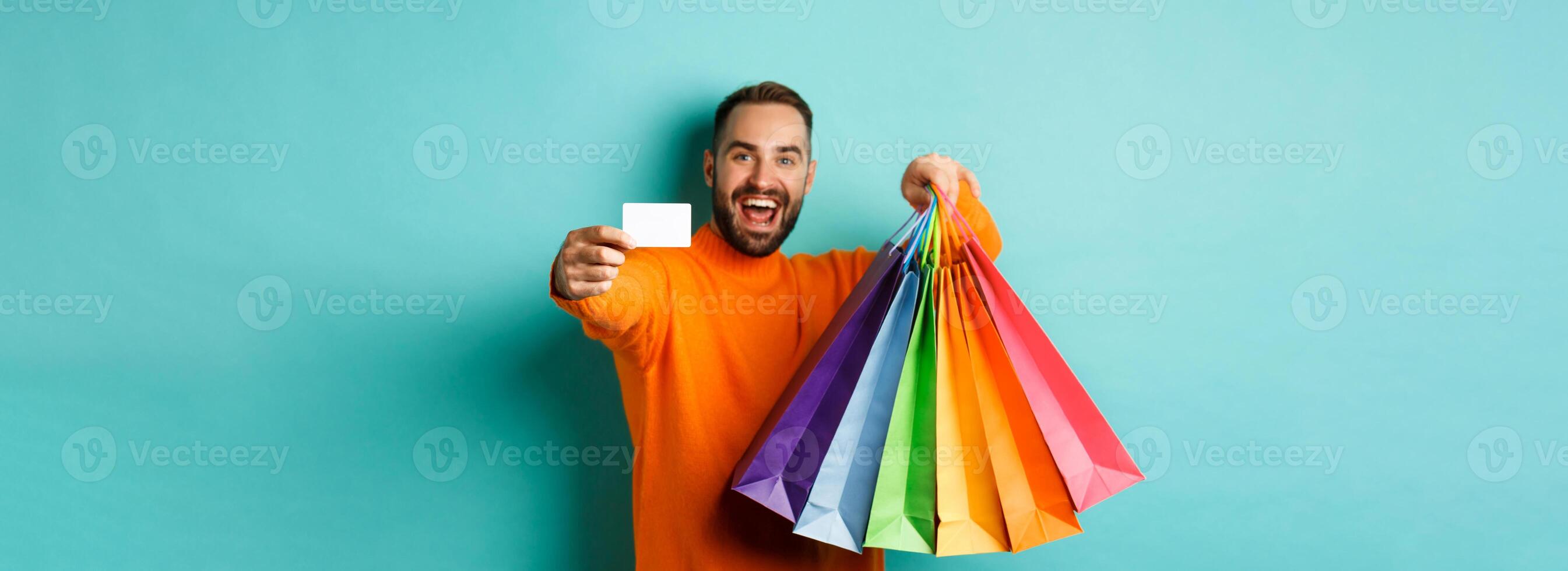 Happy aduly man showing credit card and shopping bags, standing against turquoise background photo