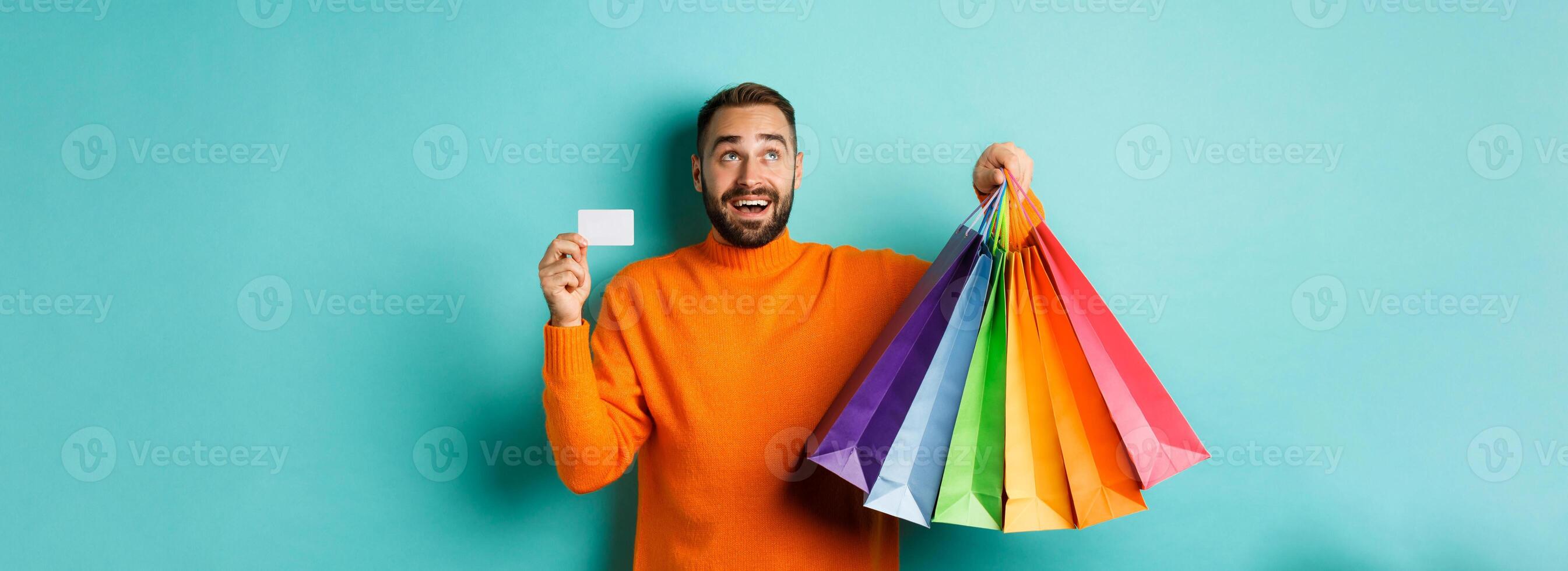Happy aduly man showing credit card and shopping bags, standing against turquoise background photo