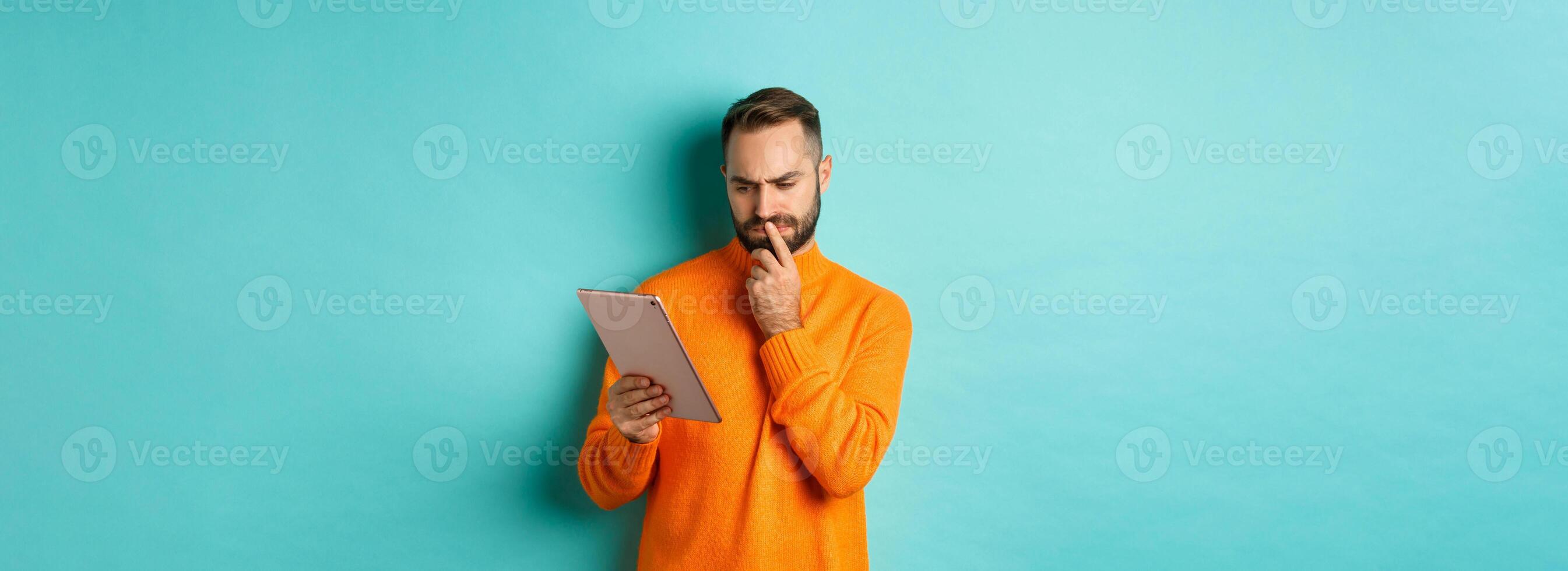 Serious man pondering and looking at digital tablet screen, reading social media, standing over turquoise background photo