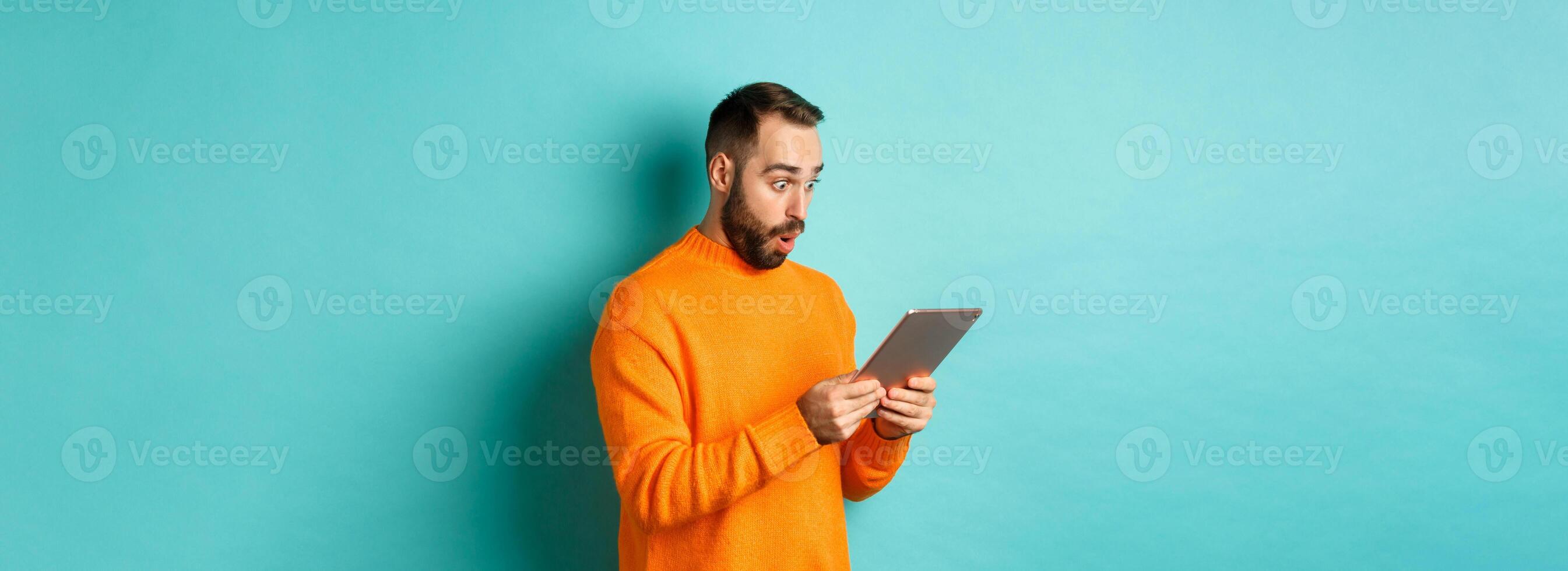 Image of male model in orange sweater staring at digital tablet screen, looking surprised, standing over light blue background photo