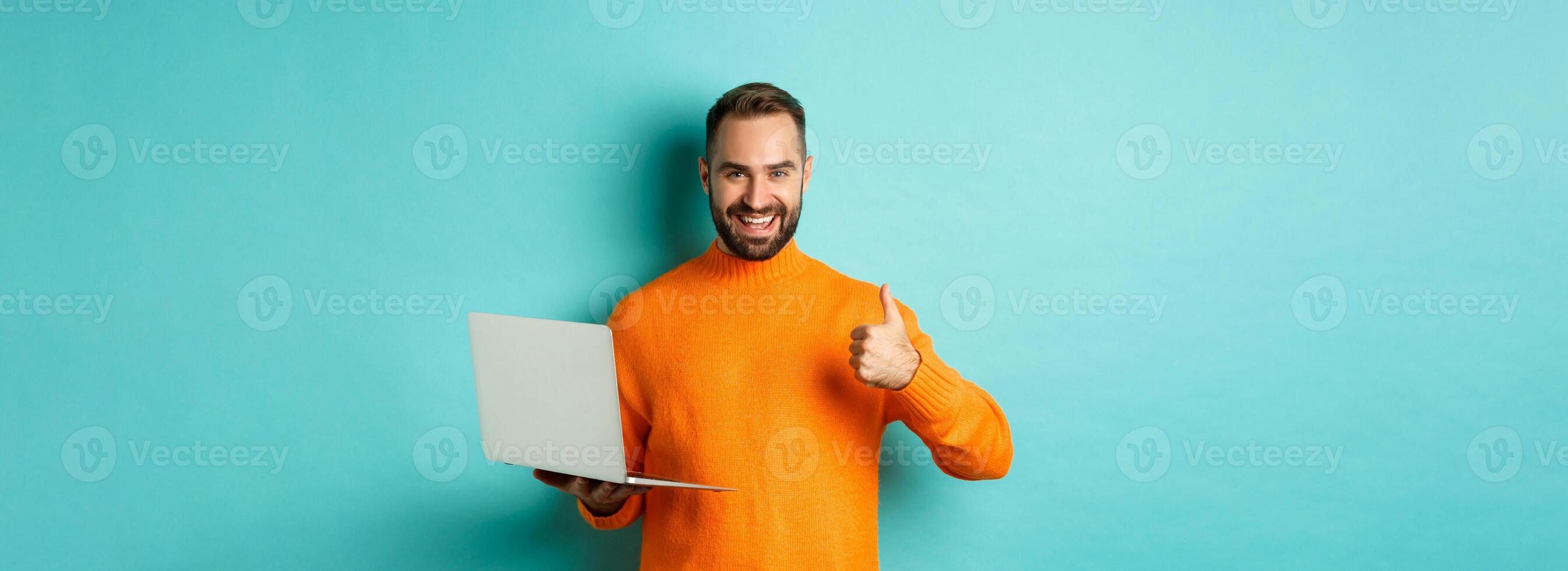 Freelance and technology concept. Lucky man in orange sweater, showing thumb up while working with laptop, standing satisfied over light blue background photo