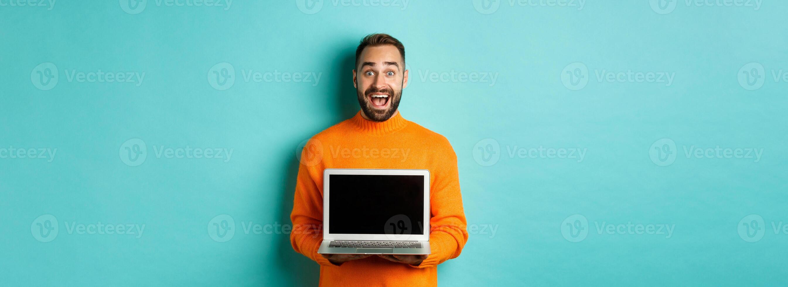 Handsome bearded man in orange sweater showing laptop screen, demonstrating promo, standing over light blue background photo
