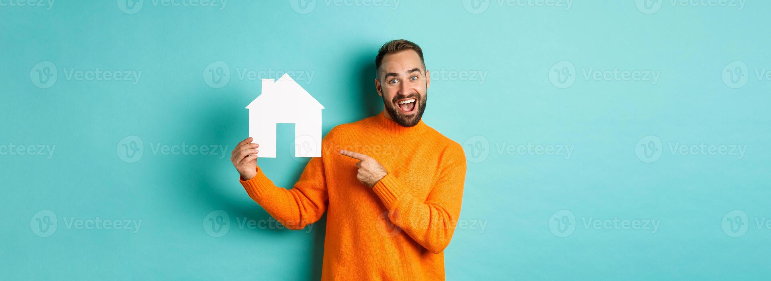 Real estate concept. Happy man pointing finger at paper home maket and smiling, standing over light blue background photo