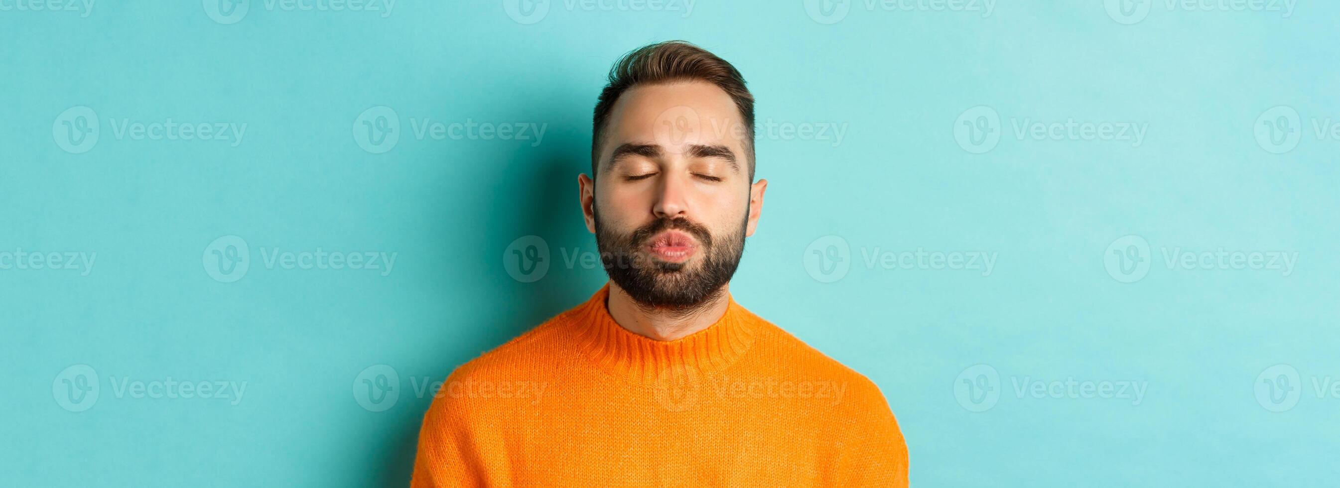 Close-up of young man in sweater pucker lips, close eyes and waiting for kiss, standing over light blue background photo