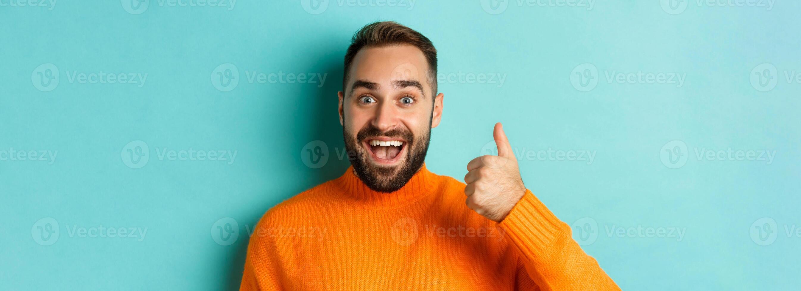 Close-up of cheerful male model showing thumb-up, smiling pleased, standing over light blue background photo