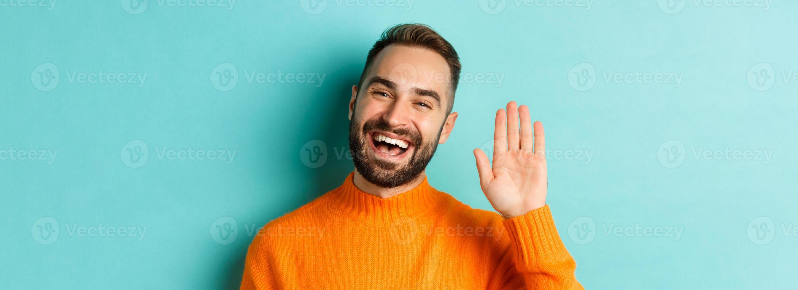 Close-up of friendly young man smiling, waving hand to say hello, greeting you, standing over light blue background photo