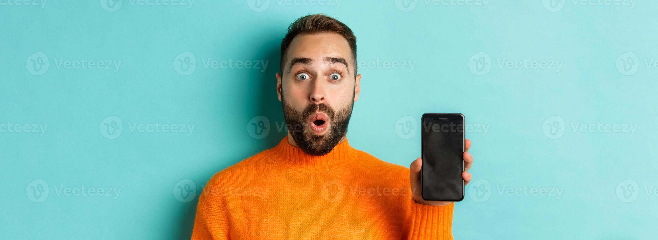 Close-up of handsome bearded guy in orange sweater, showing smartphone screen and smiling, showing promo online, turquoise background photo