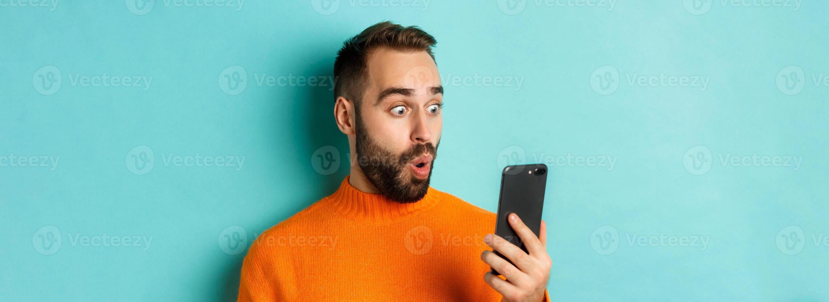 Close-up of caucasian man staring at phone screen with surprised face, wearing orange sweater, light blue background photo