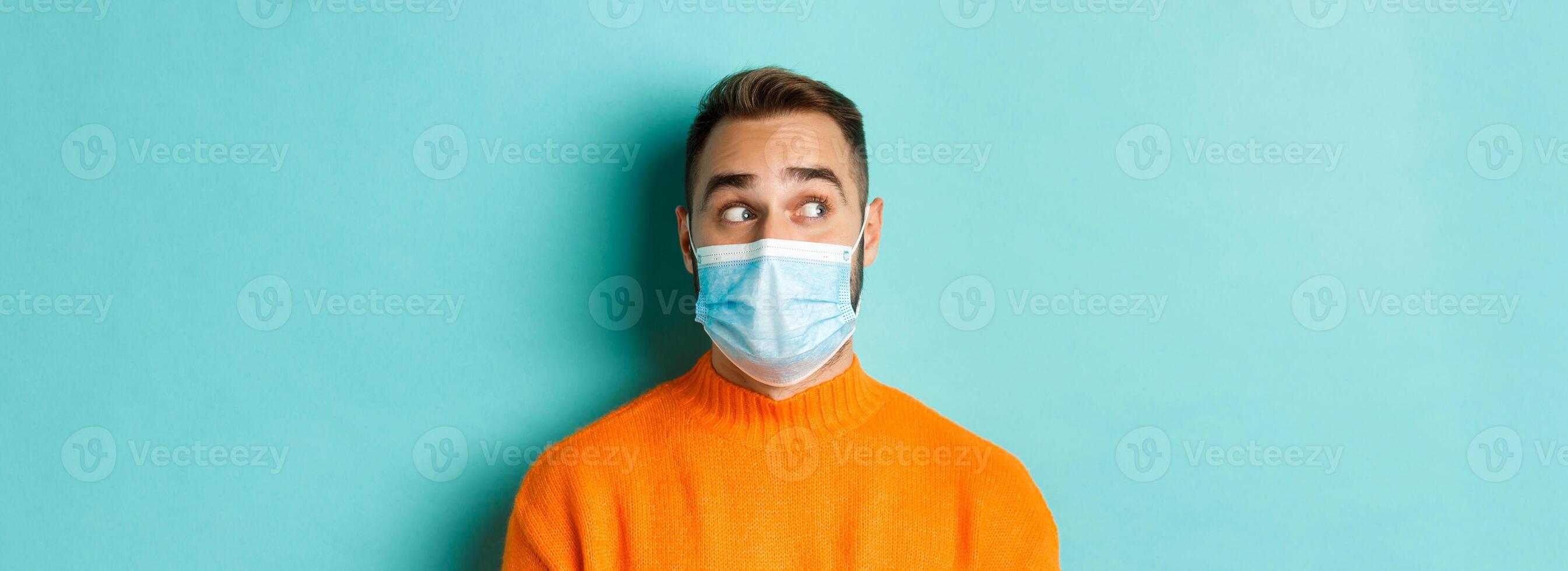 Covid-19, social distancing and quarantine concept. Close-up of young male model in medical mask looking left curious, staring at promo copy space, light blue background photo