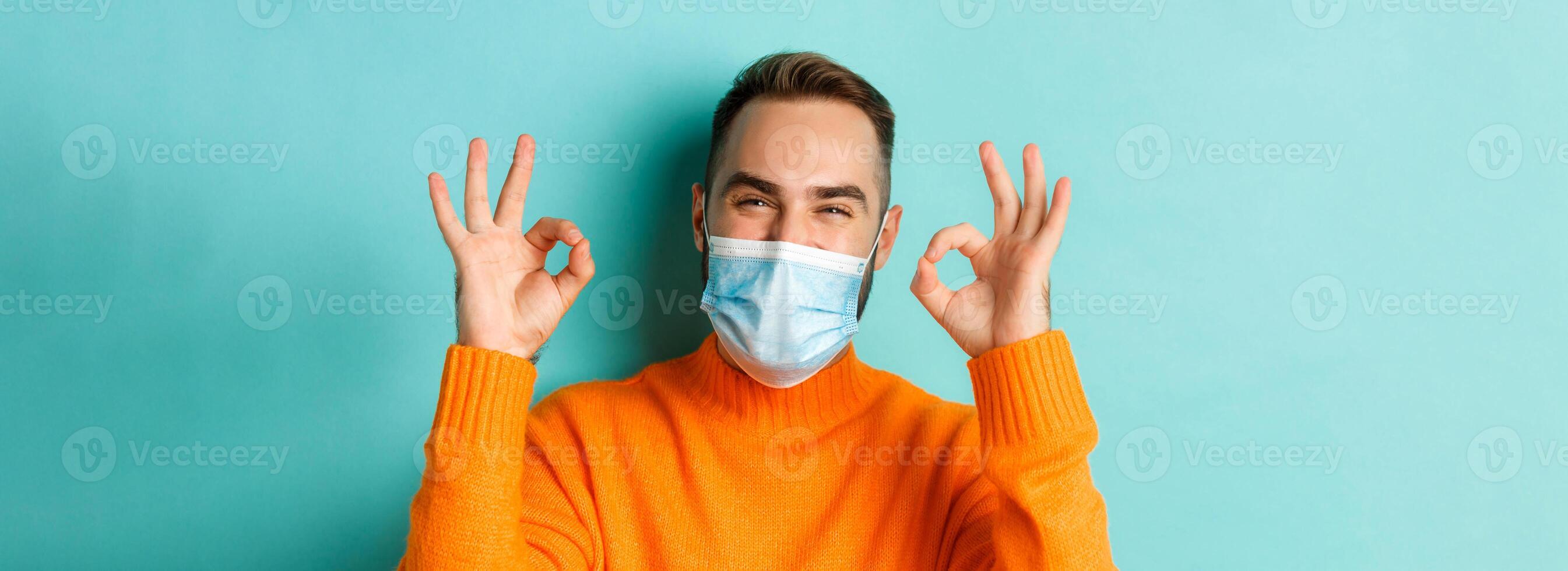 Covid-19, social distancing and quarantine concept. Close-up of satisfied male model in medical mask showing okay signs, praising and agreeing, standing over light blue background photo