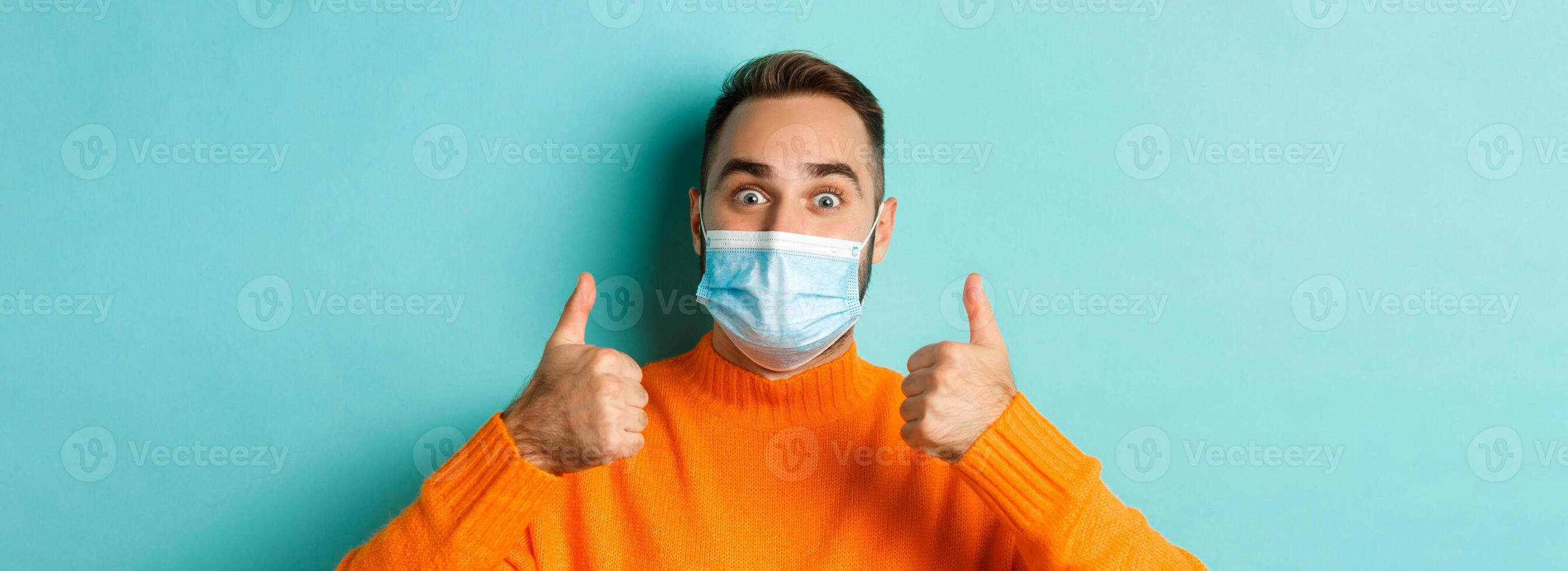 Covid-19, social distancing and quarantine concept. Close-up of satisfied male model in medical mask showing thumbs-up, praising and agreeing, standing over light blue background photo