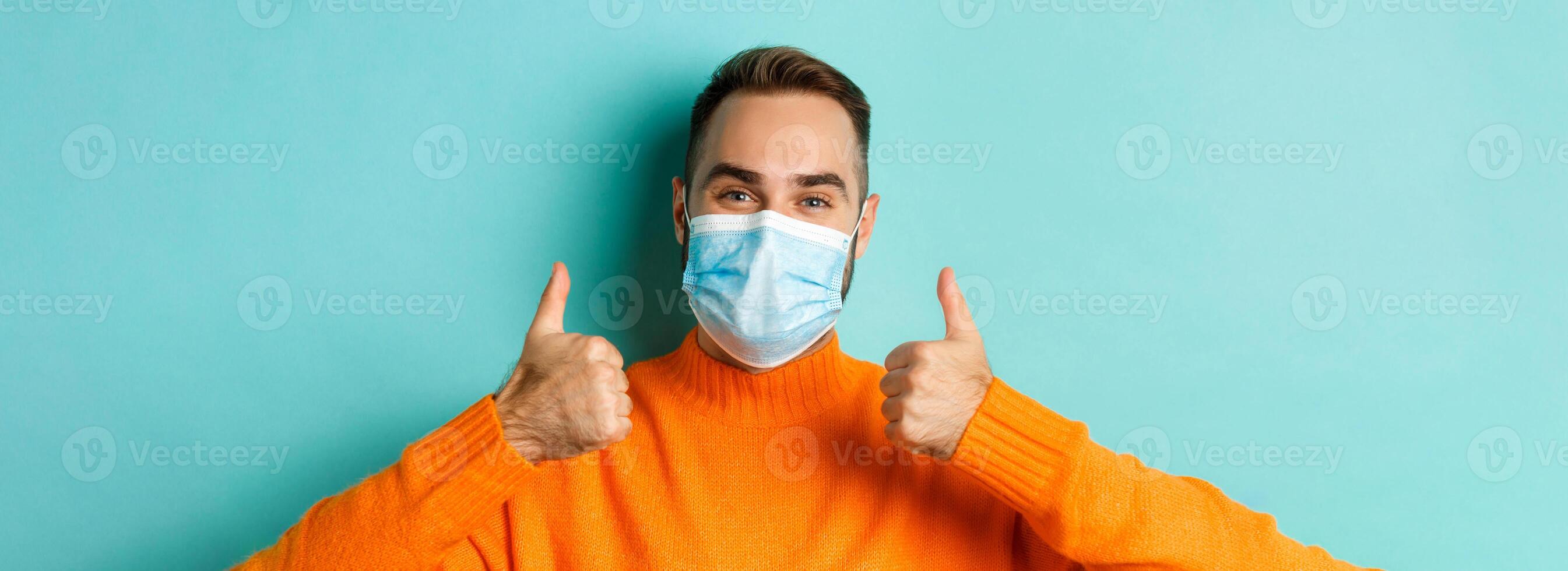 Covid-19, social distancing and quarantine concept. Close-up of satisfied male model in medical mask showing thumbs-up, praising and agreeing, standing over light blue background photo