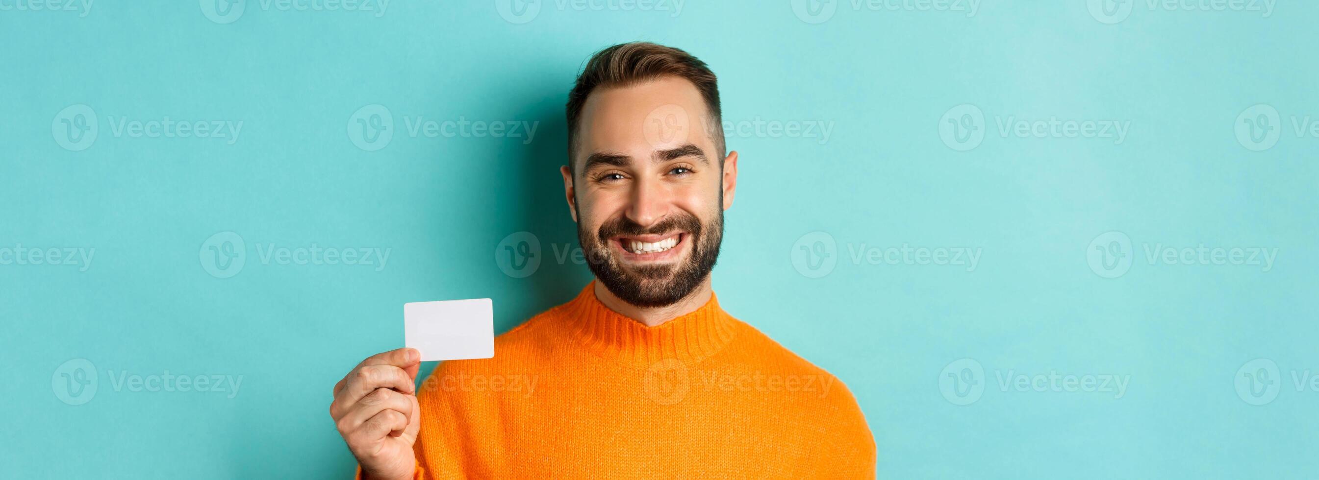 Close-up of handsome caucasian man going on shopping, showing credit card and smiling, standing over turquoise background photo