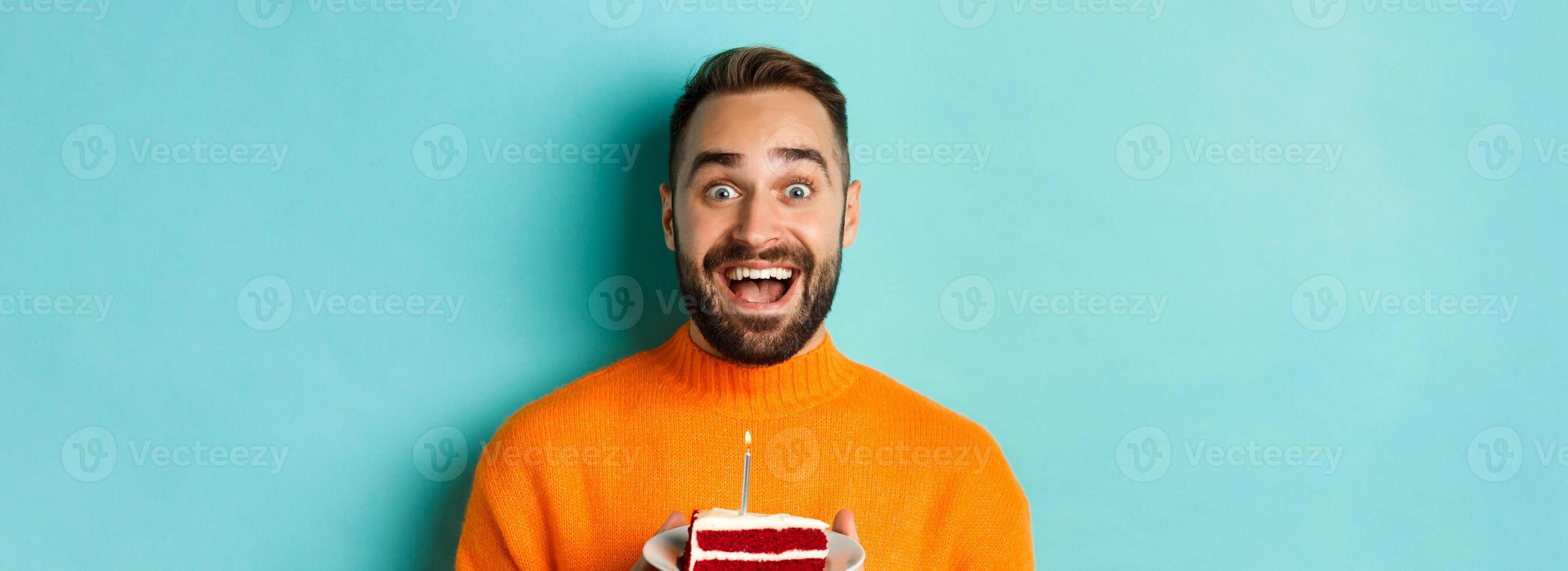 Close-up of happy adult man celebrating birthday, holding bday cake with candle and making wish, standing against turquoise background photo