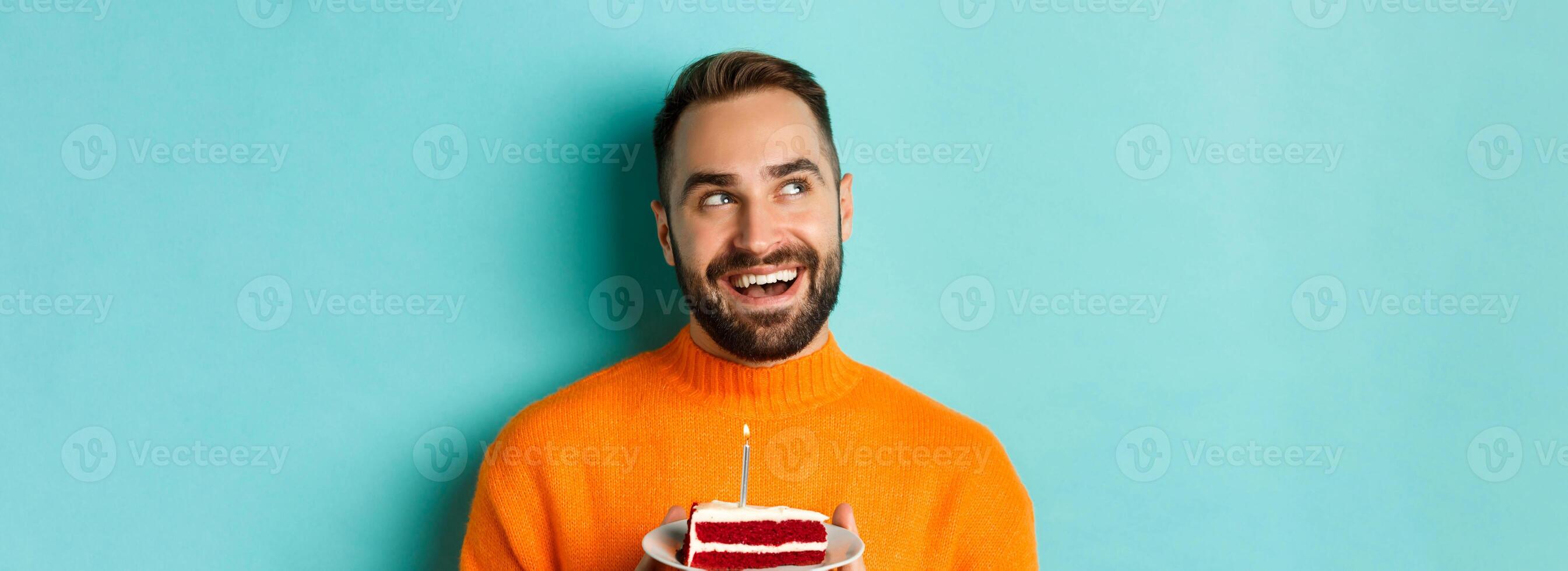 Close-up of happy adult man celebrating birthday, holding bday cake with candle and making wish, standing against turquoise background photo