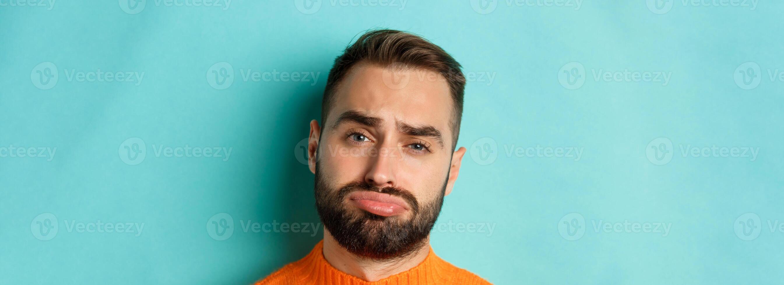 Headshot of sad and gloomy man complaining, pouting and frowning disappointed, standing against turquoise background photo