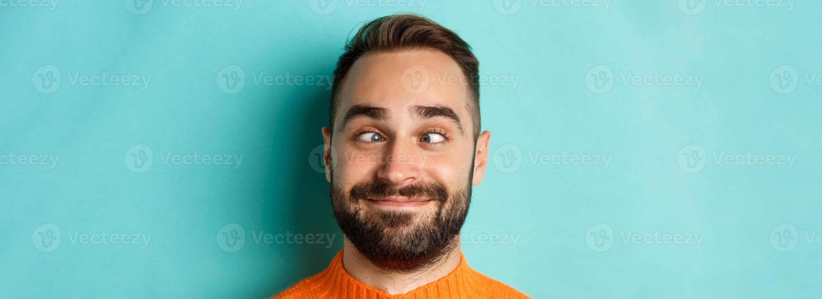 Headshot of funny caucasian man making faces, squinting and smiling silly, standing over light blue background photo