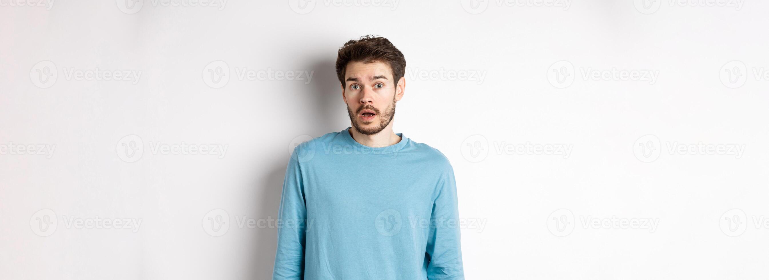 Confused caucasian guy drop jaw and stare startled at camera, standing over white background photo