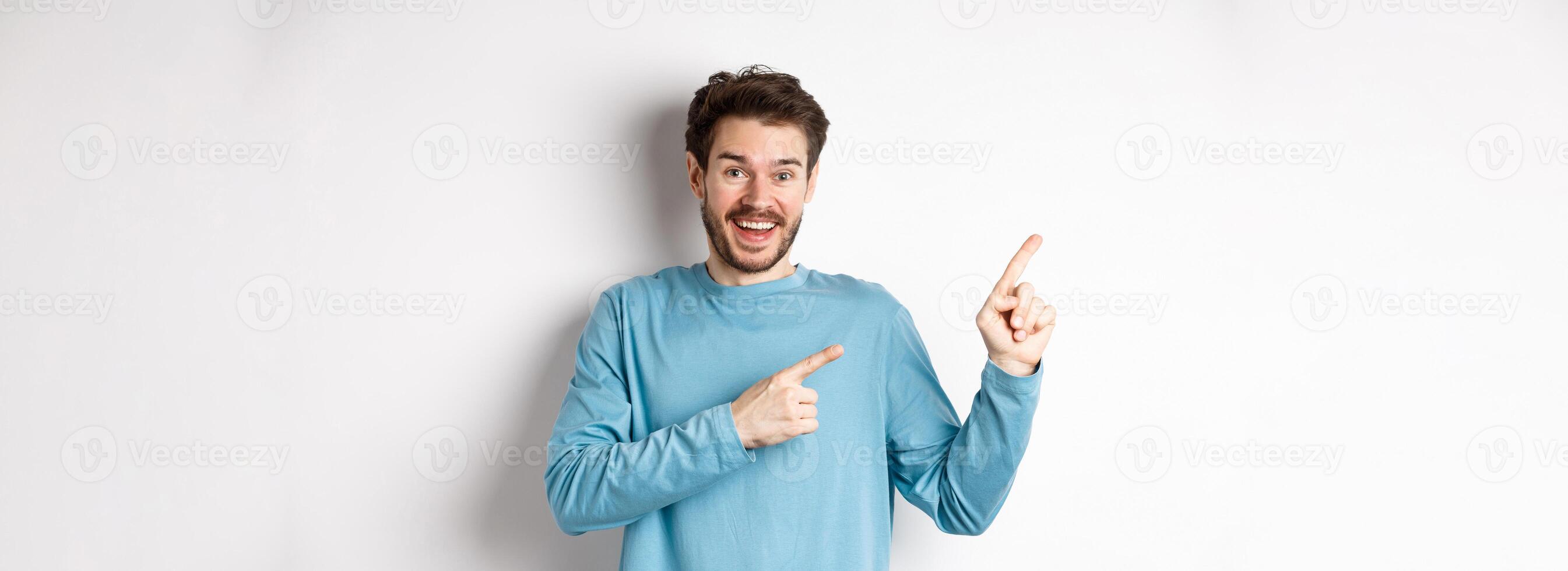 Happy caucasian man in casual blue sweatshirt, pointing fingers at upper left corner, showing link or logo on white background, smiling at camera photo