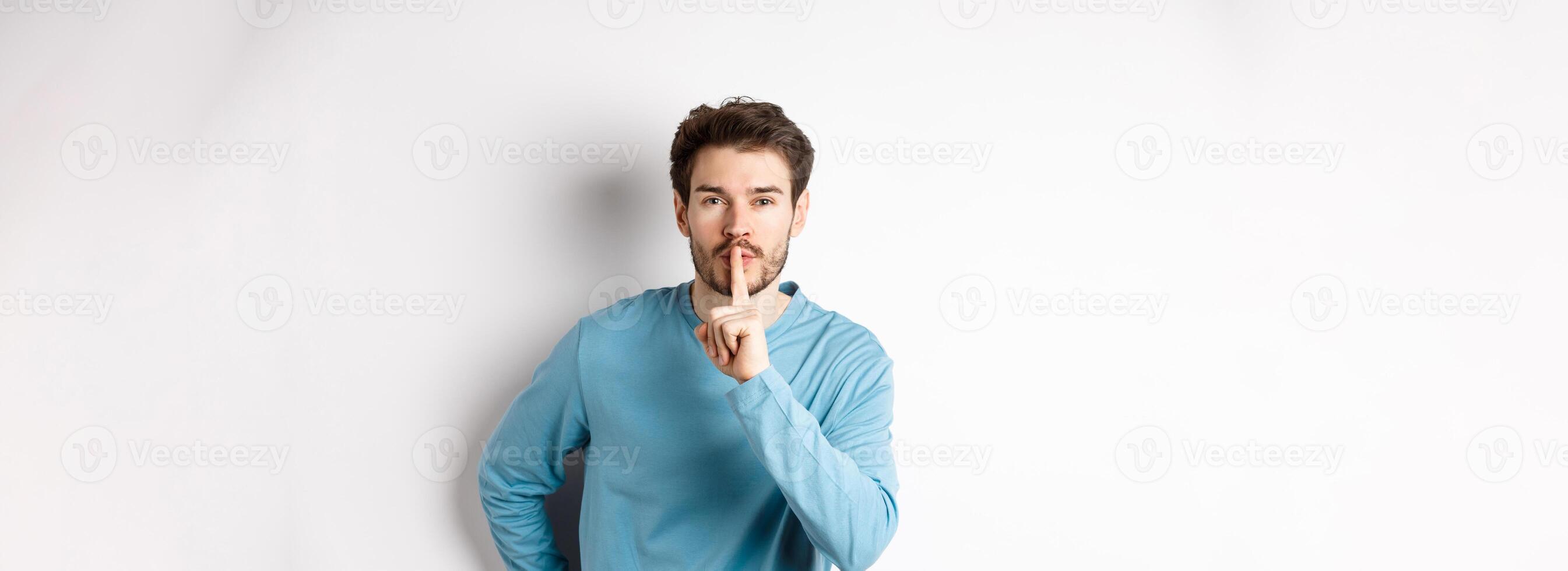 Attractive bearded man asking to keep quiet, showing taboo hush gesture and looking at camera calm, standing over white background photo