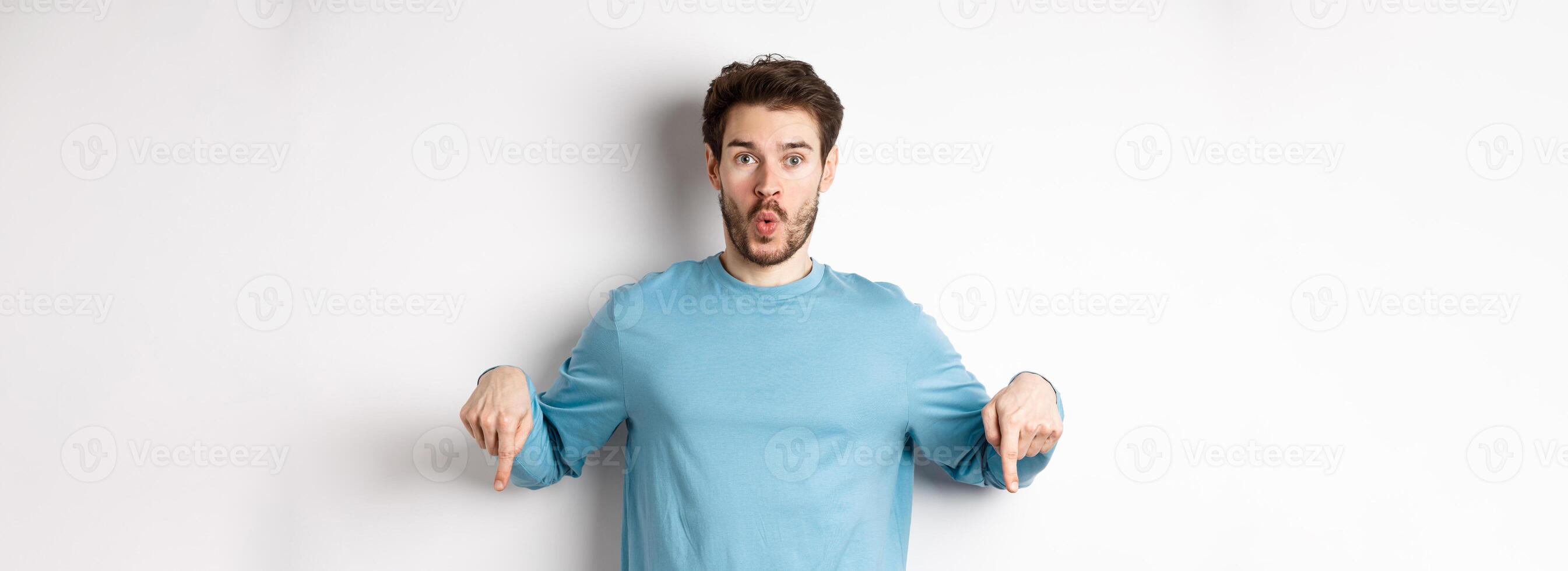Amazed young man with beard, standing in casual blue shirt, saying wow and pointing down, look here gesture, standing over white background photo