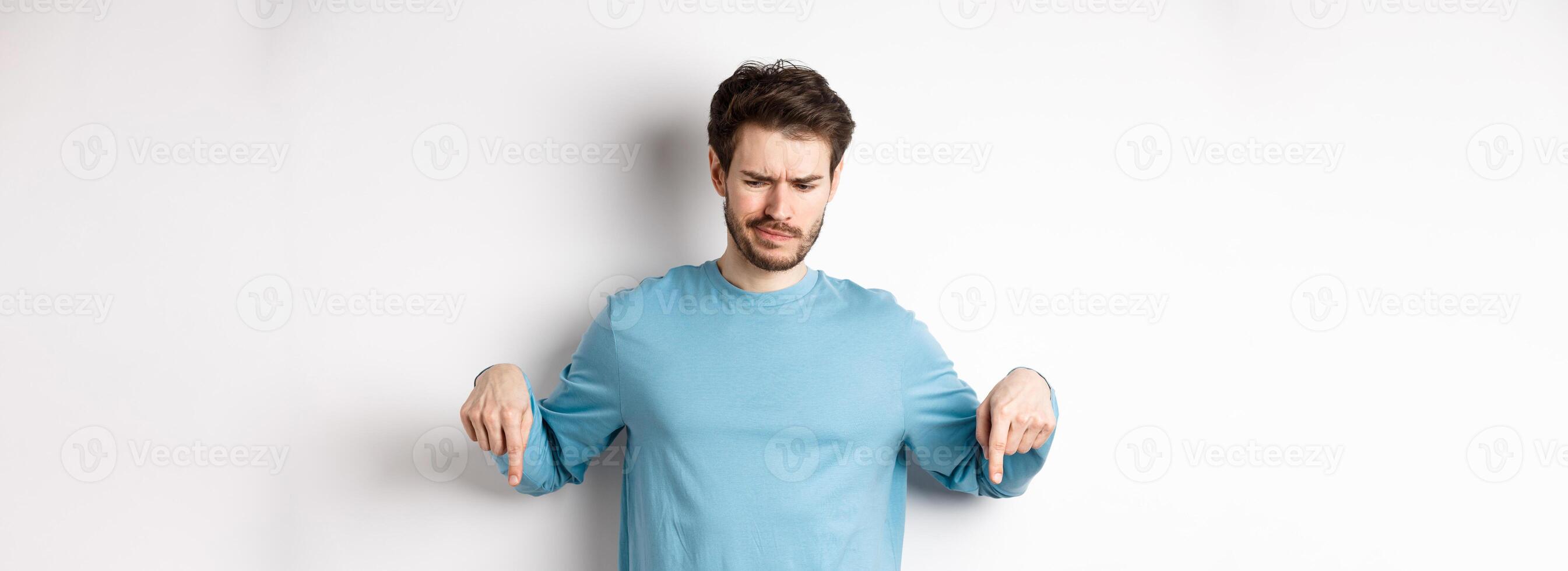 Skeptical boy pointing fingers down and frowning disappointed, looking at bad promo offer, standing on white background photo
