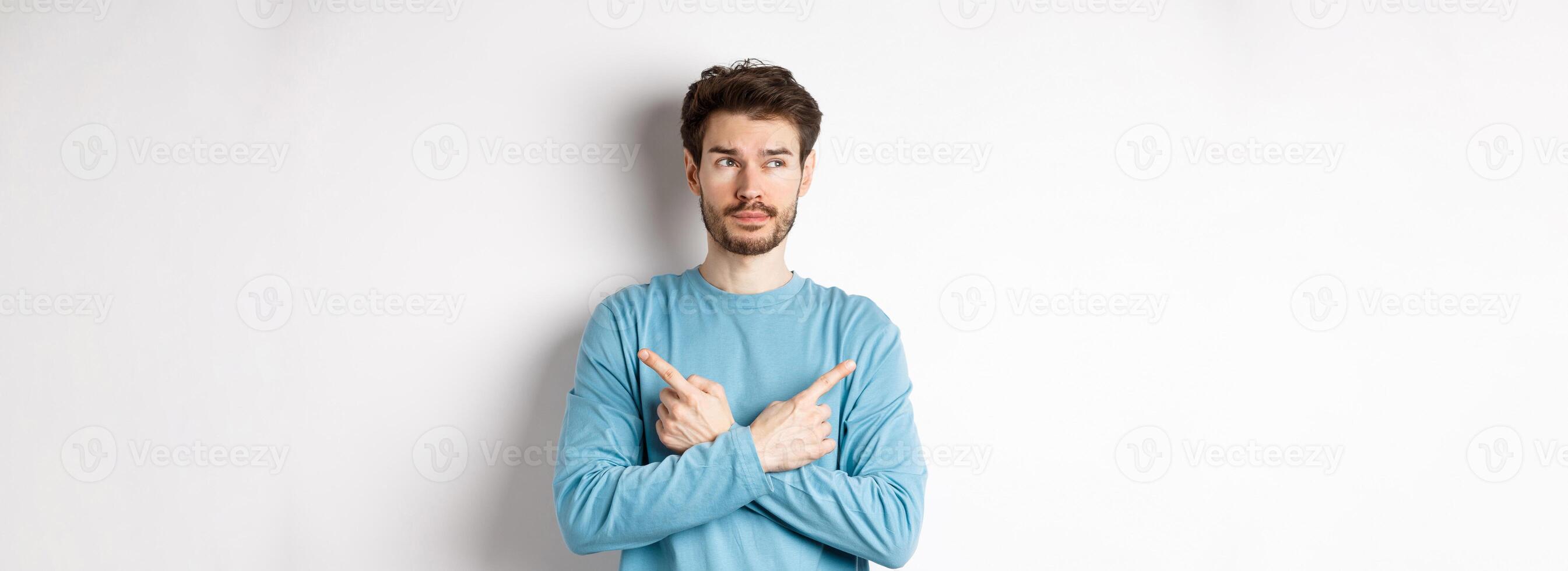 Indecisive man making choice, pointing fingers sideways at two variants and deciding, looking pensive at left side, standing on white background photo